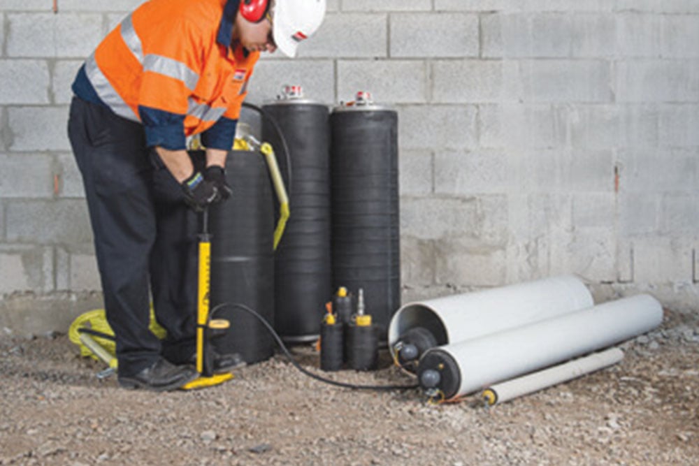 A worker in a high-visibility orange jacket and safety gear uses a manual pump to inflate a black pipe plug. Several other pipe plugs and related equipment are placed on the ground near a concrete block wall.