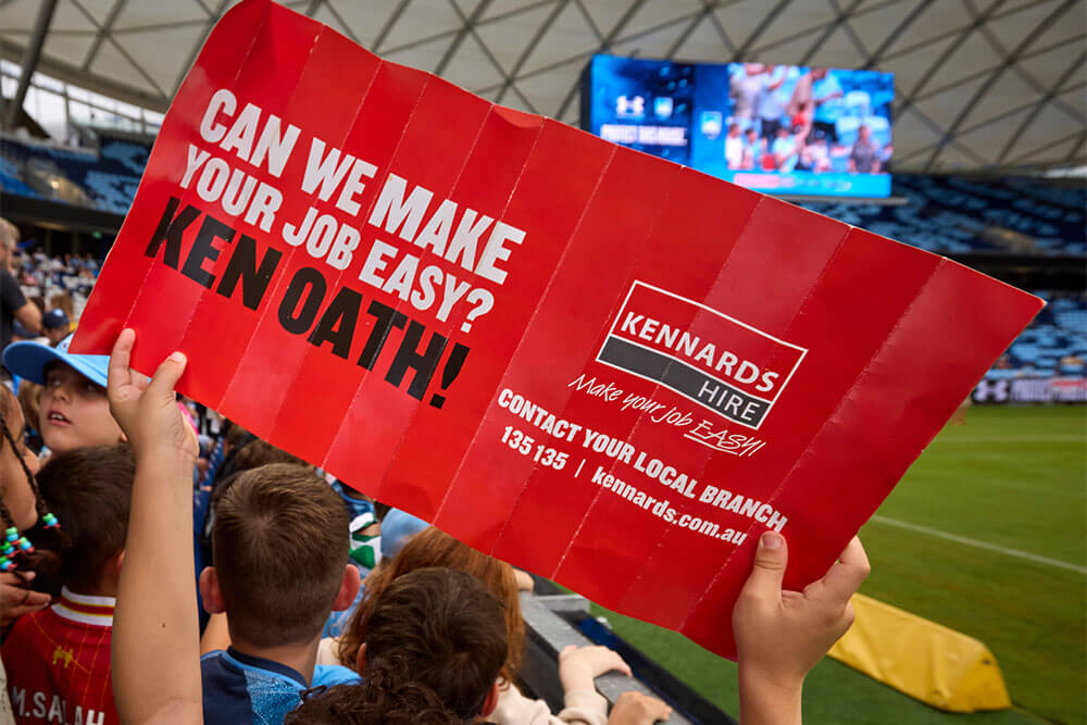Sydney FC fans holding up a Kennards Hire sign in the stands